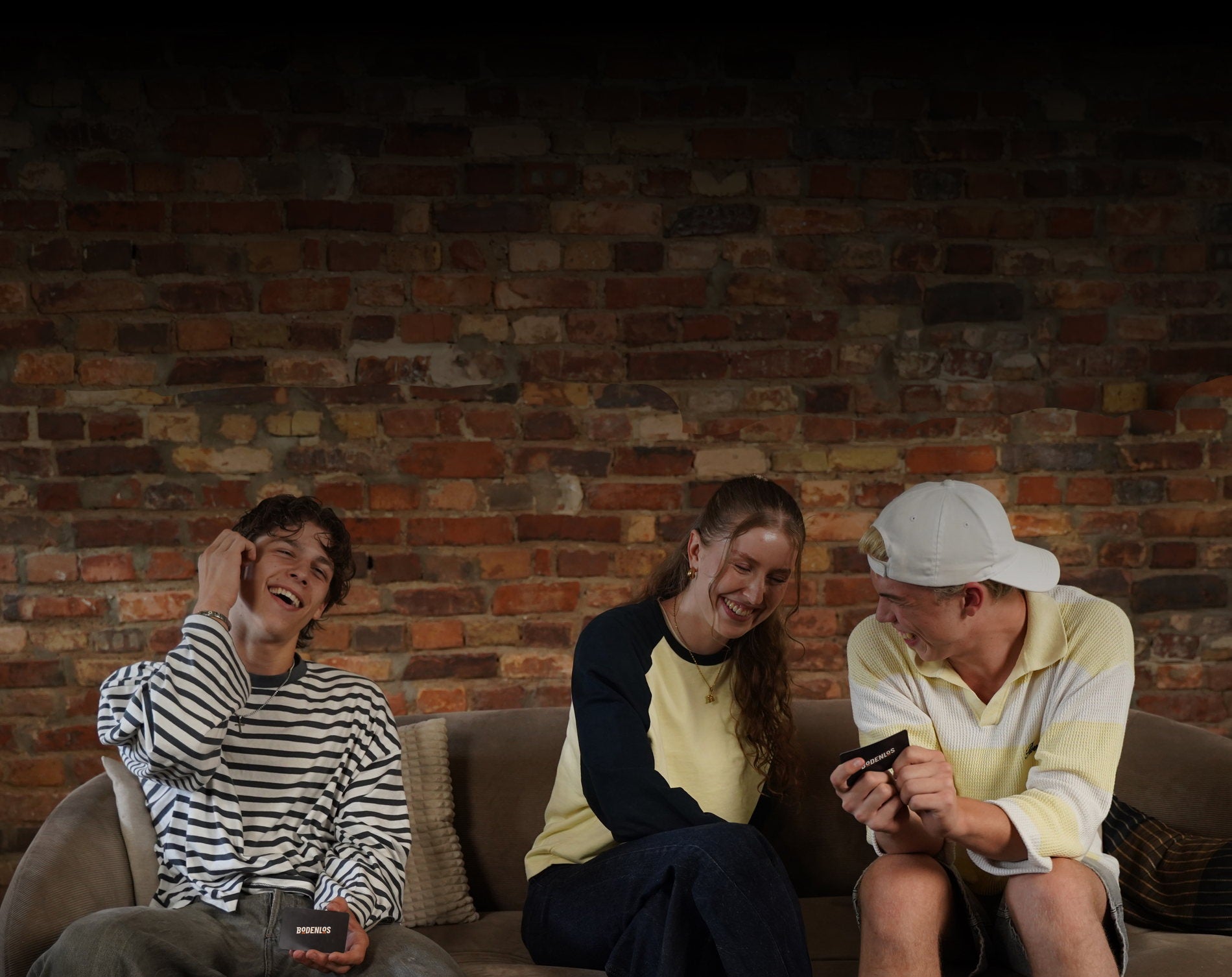 Drei junge Leute lachen auf dem Sofa vor einer Ziegelwand